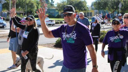 A group of people walking outdoors in a community event, wearing matching purple shirts that read “Together to Fight Suicide,” some waving toward the camera.