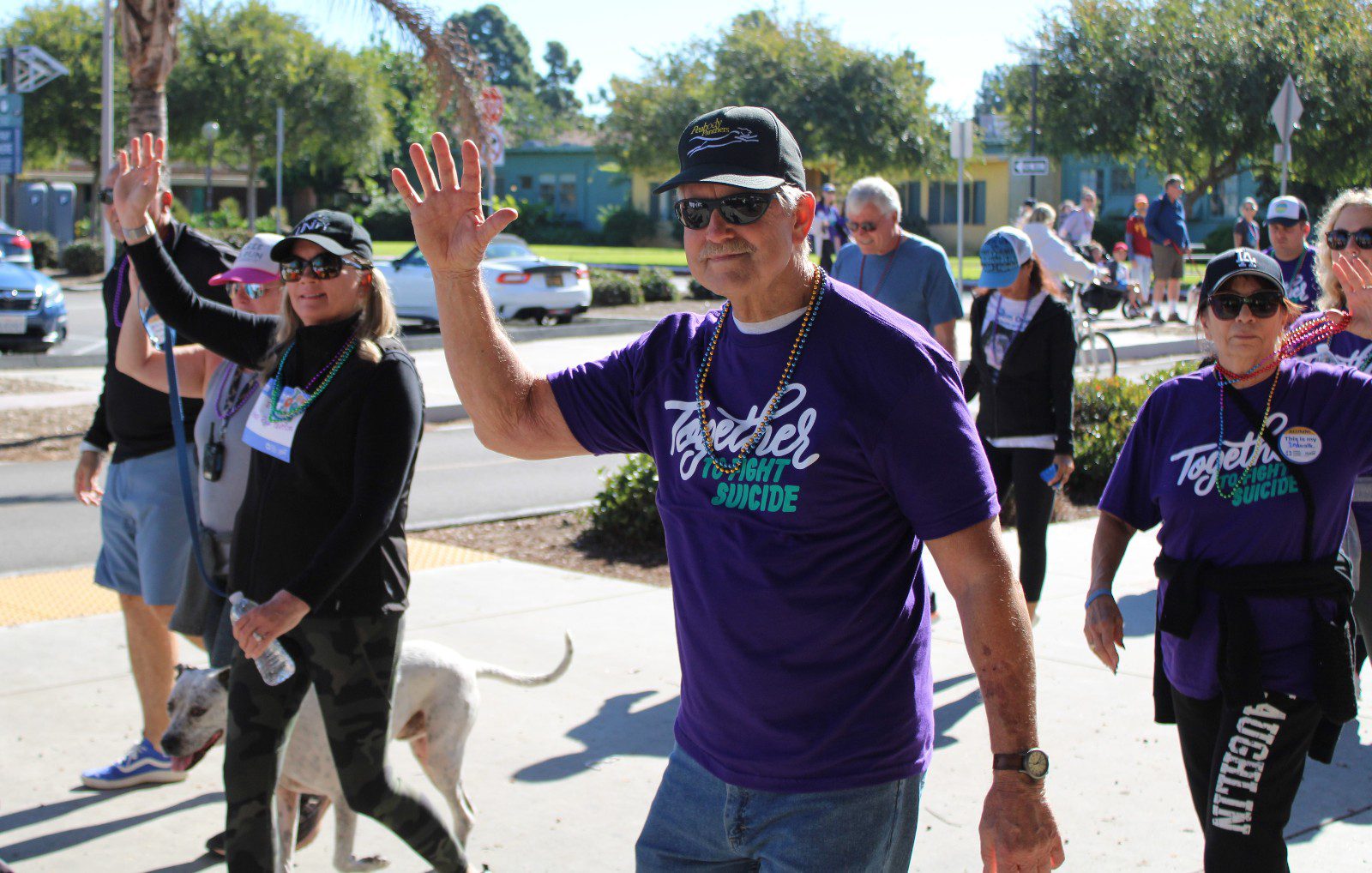 A group of people walking outdoors in a community event, wearing matching purple shirts that read “Together to Fight Suicide,” some waving toward the camera.