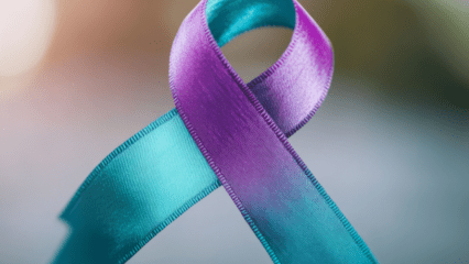 The image shows a single awareness ribbon made of satin material. The ribbon features two distinct colors: one half is teal and the other half is purple. The ribbon is looped in the traditional awareness‑ribbon shape, with the ends crossing at the bottom and the top forming a smooth rounded loop. The texture of the ribbon appears slightly glossy, reflecting soft light that highlights subtle folds and stitching along the edges. The background is blurred with muted tones, creating a gentle gradient that keeps focus on the ribbon itself. No text or additional objects are present in the image.