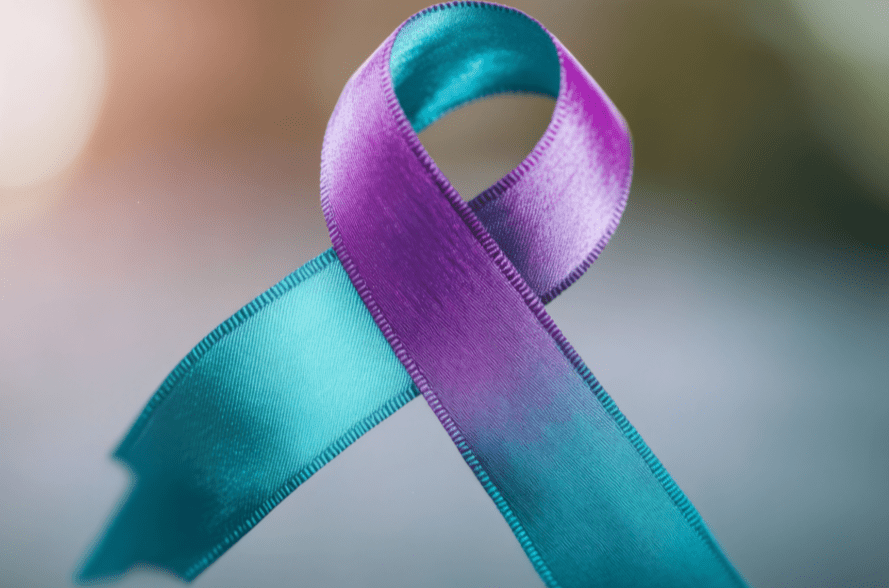 The image shows a single awareness ribbon made of satin material. The ribbon features two distinct colors: one half is teal and the other half is purple. The ribbon is looped in the traditional awareness‑ribbon shape, with the ends crossing at the bottom and the top forming a smooth rounded loop. The texture of the ribbon appears slightly glossy, reflecting soft light that highlights subtle folds and stitching along the edges. The background is blurred with muted tones, creating a gentle gradient that keeps focus on the ribbon itself. No text or additional objects are present in the image.
