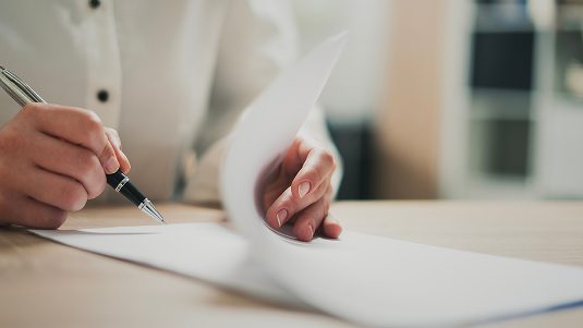 A person at a table with papers, appearing to sign the last page.