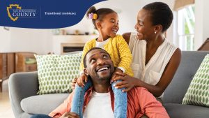 A happy family sits together in a bright living room. A young girl in a yellow sweater sits on her father’s shoulders while he looks up at her, smiling. The mother, sitting beside them on the couch, leans in with a big smile as she holds her daughter’s arm.