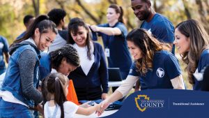 Smiling volunteers in matching blue shirts greet families at an outdoor community event. A young girl shakes hands with a friendly volunteer across a table with informational materials, lanyards, and folded shirts.
