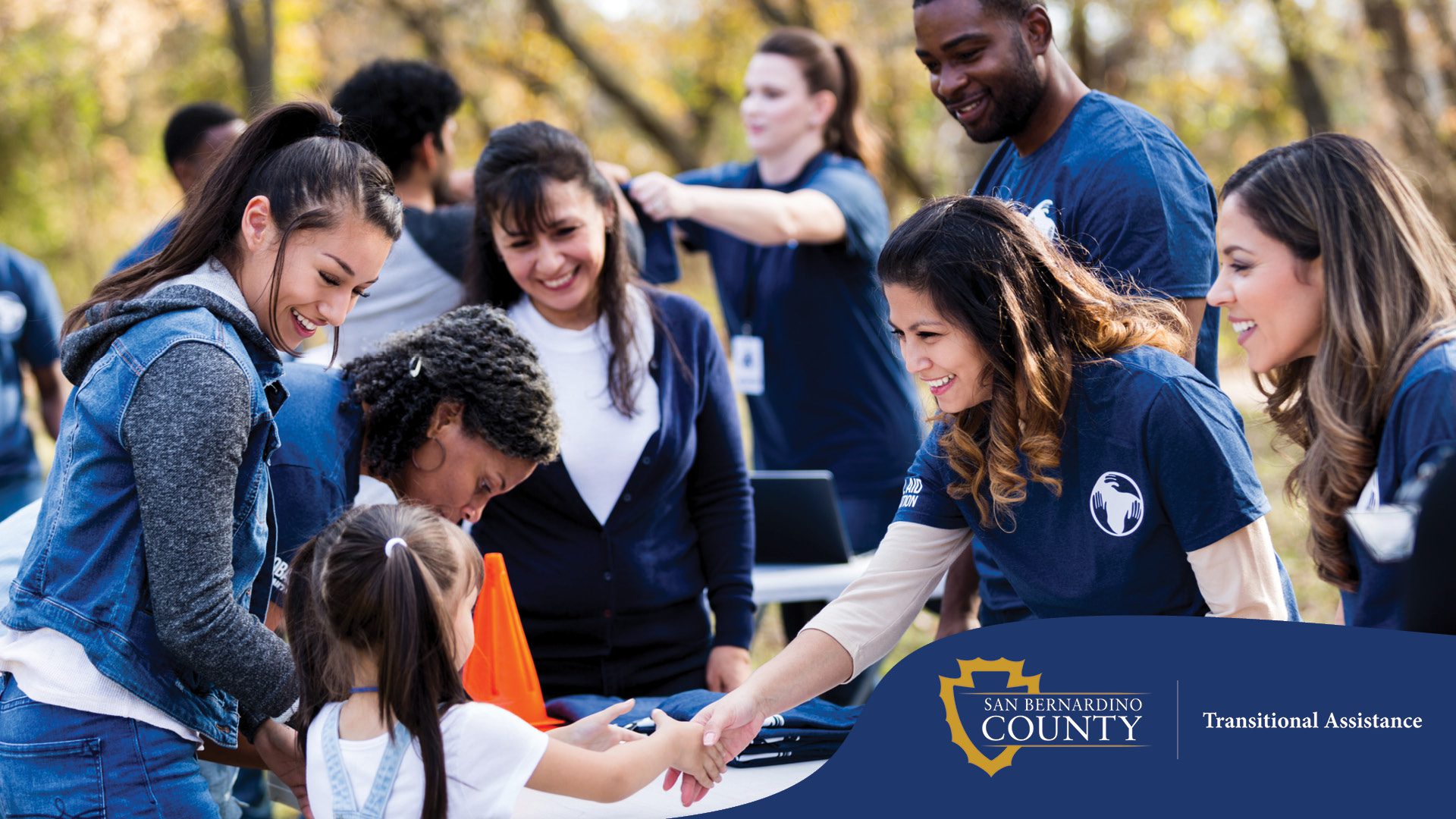 Smiling volunteers in matching blue shirts greet families at an outdoor community event. A young girl shakes hands with a friendly volunteer across a table with informational materials, lanyards, and folded shirts.