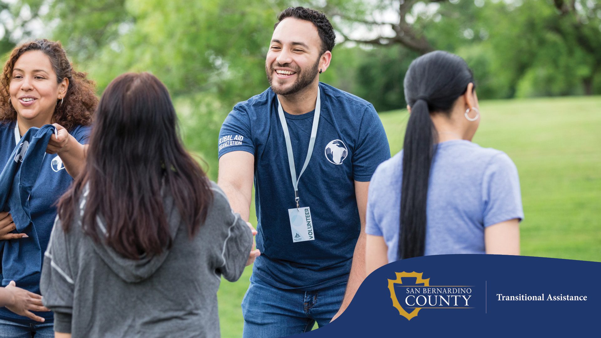 A male volunteer smiles warmly as he reaches out to shake hands with an attendee at an outdoor event. Other volunteers stand beside him, handing out t-shirts. Everyone is gathered around a table set up on a grassy field with trees in the background, creating a friendly and community-focused environment.