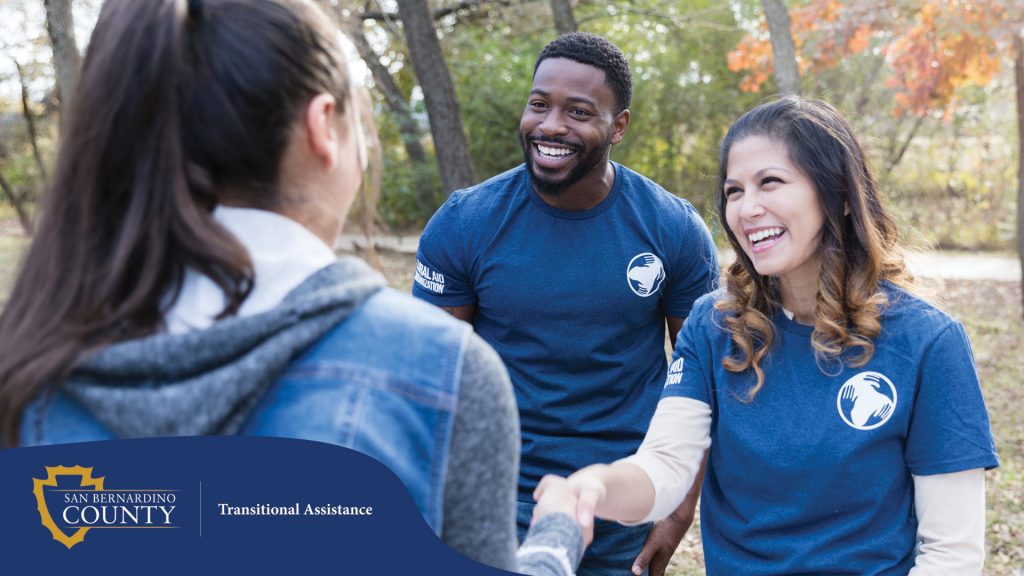 A smiling woman and man, both wearing blue volunteer t-shirts, greet and shake hands with a visitor outdoors at a community event.