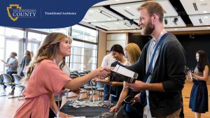 A smiling woman at a check-in table shakes hands with a man holding an event program and wearing a name badge. Behind them, several other attendees are checking in or reviewing materials.