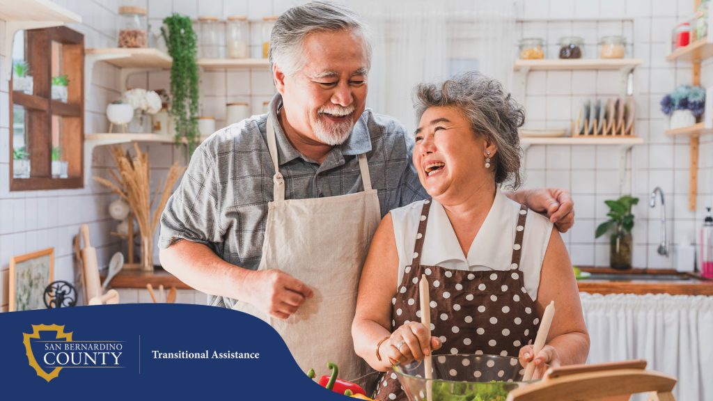 Older couple laughing while preparing a salad together in a bright kitchen, surrounded by fresh vegetables.