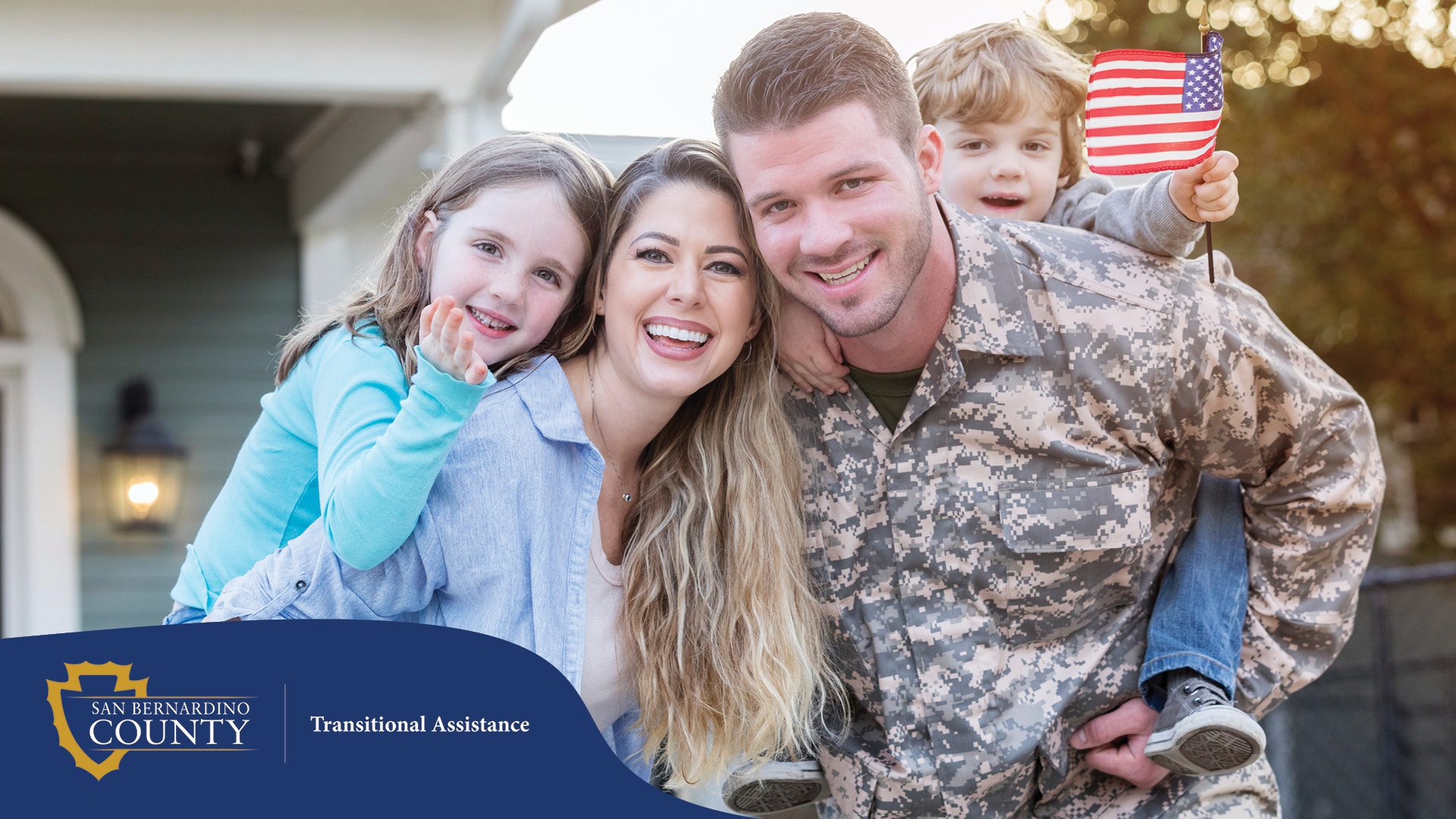 A smiling family enjoys time together outdoors. A woman carries a young girl on her back, while a man in military camouflage uniform carries a young boy holding a small American flag.