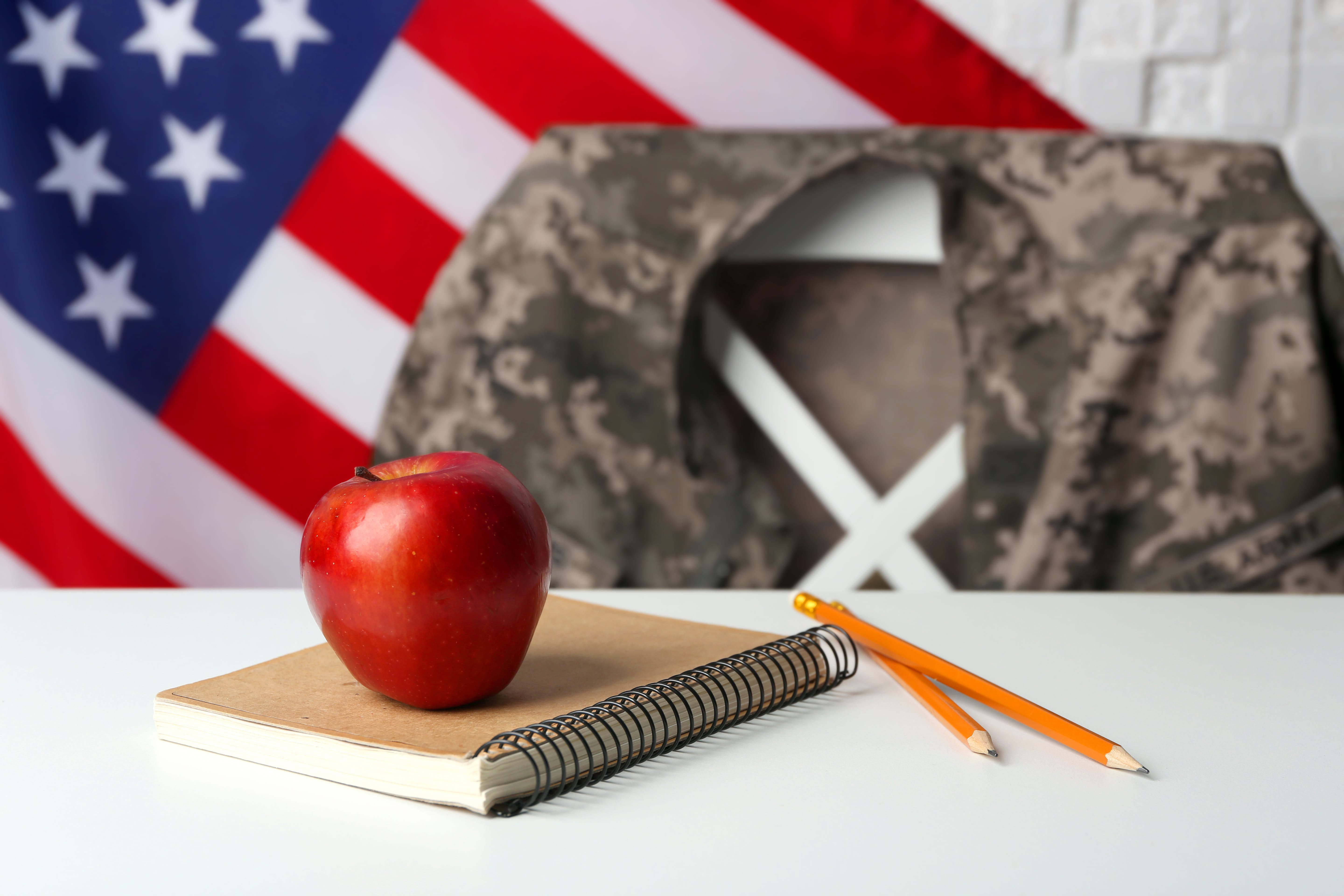 A notebook with an apple on top of it and 2 pencils. An American flag in the background with an Army Jacket draped over a chair.